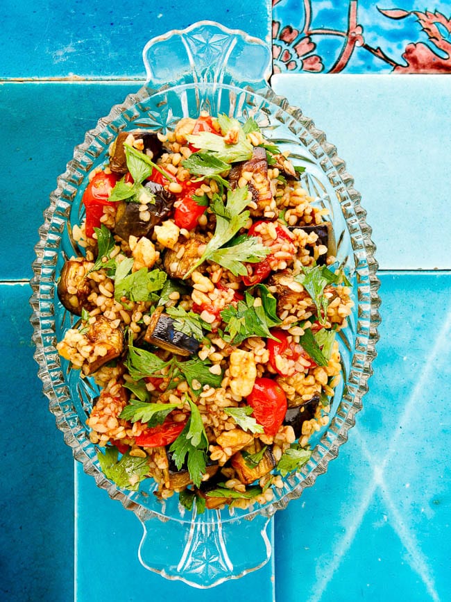 Bulgur salad with tomato and aubergine in transparent dish on turqoise tiles seen from above