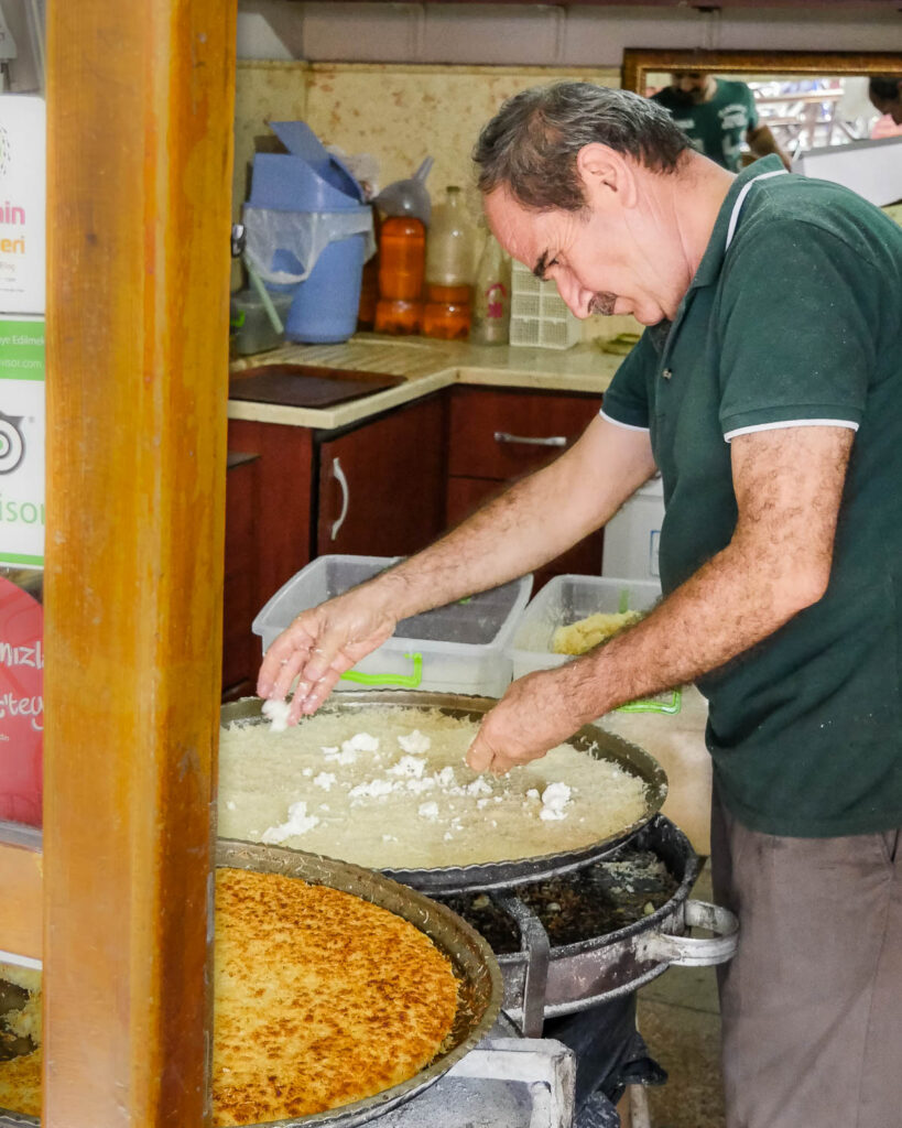 A middle aged man makes künefe by placeing cheese onto the bottom layer of kataif