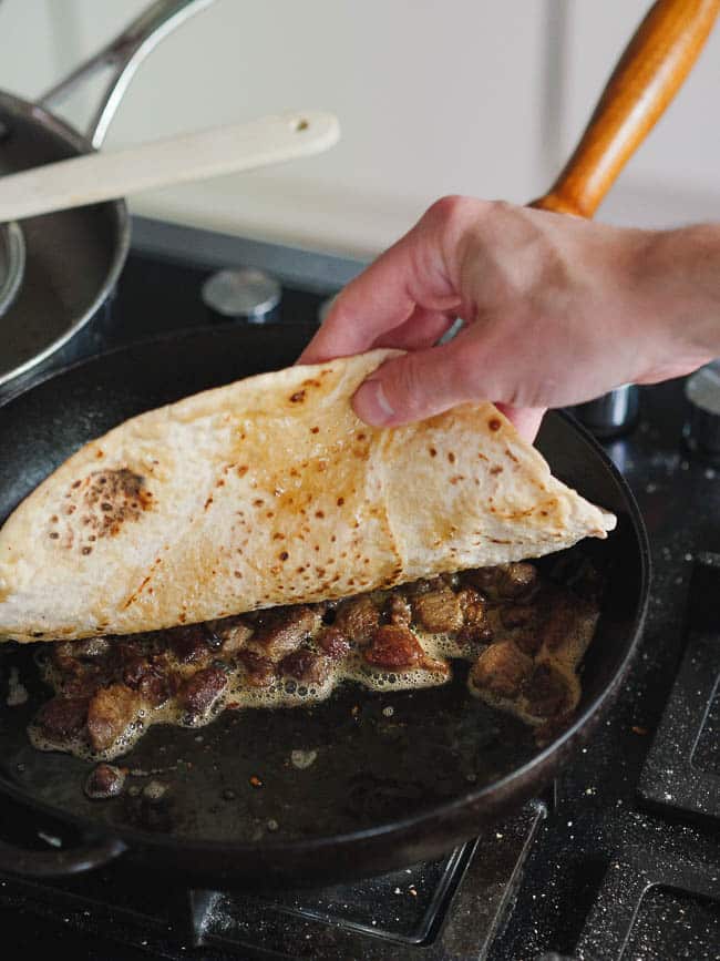 Hand lifting a lavash wrap bread from on top of the meat in frying pan