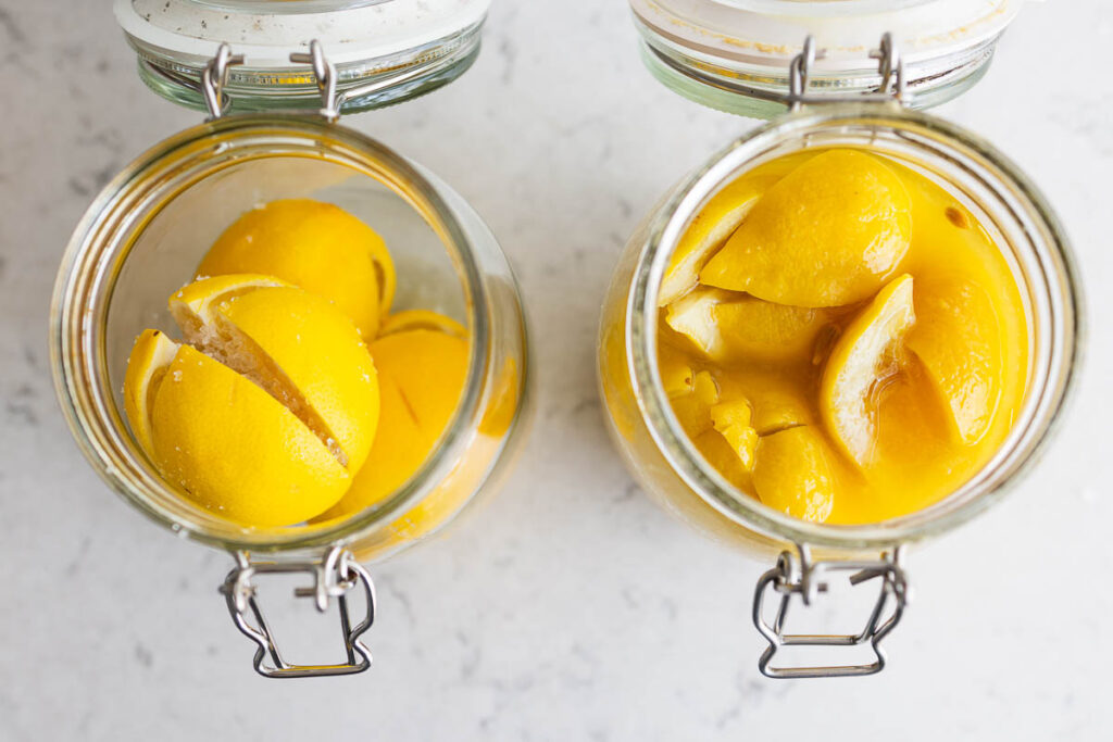 Homemade preserved lemons in jars on marble countertop, seen from above, with freshly made ones on the left and ready ones on the right