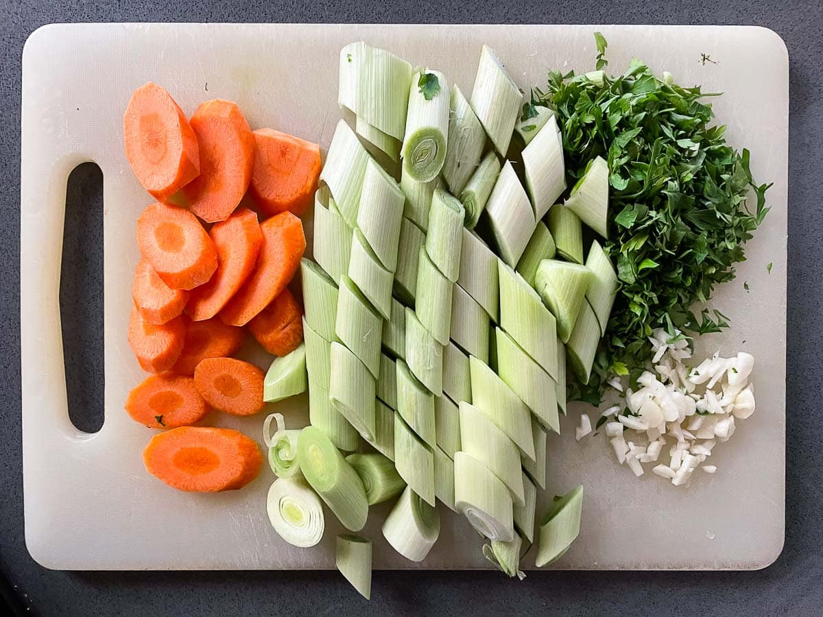 Chopped carrots, leeks, herbs and garlic on a white plastic chopping board, seen from above