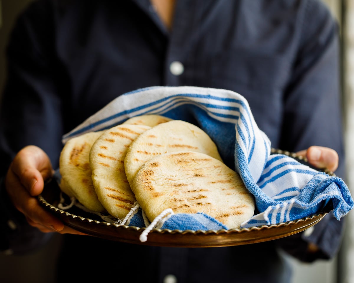 Pita bread on a tray held buy a main wearing a dark shirt. The angle is almost horizontal to the pita breads, which are wrapped in a Turkish blue and white cloth.