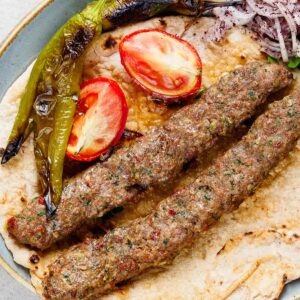 Close-up image of two Adana kebabs, two pieces of grilled tomato and two grilled long green peppers on a lavash bread. An onion salad also on the plate is barely visible in the top right corner.
