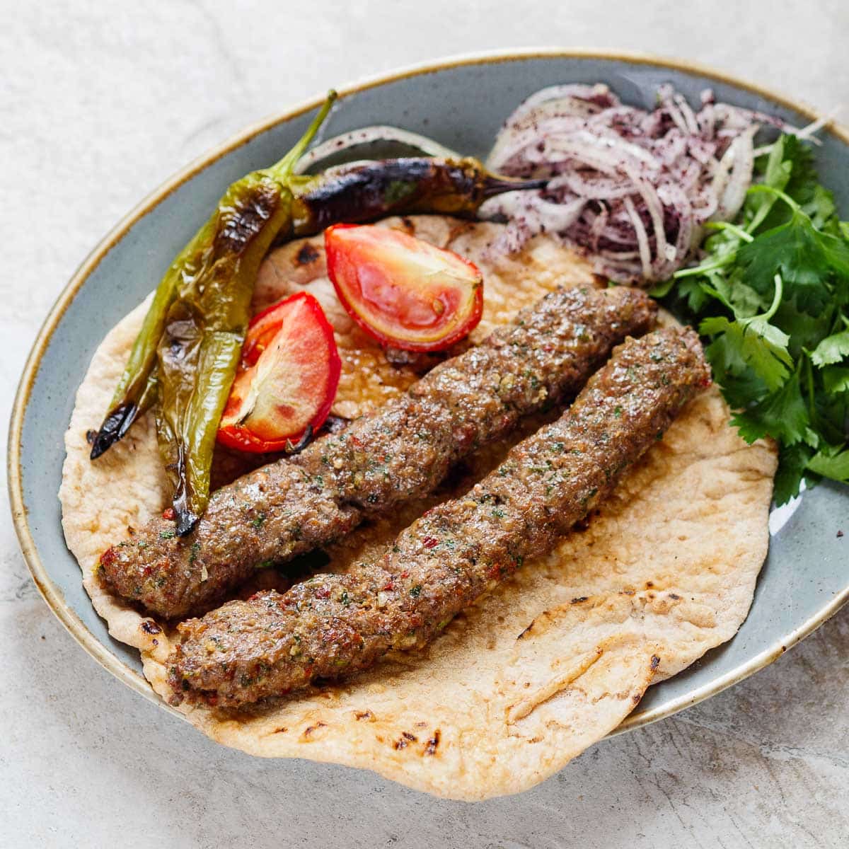 Two Adana kebabs, two pieces of grilled tomato and two grilled long green peppers on a lavash bread. There's also onion salad and parsley on the oval plate set on a light stone background. Seen from eye level