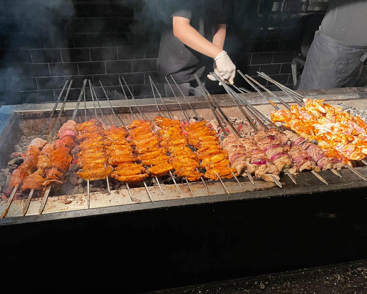Close up image of a huge number of skewers with various types of meat on them set on a very hot Turkish barbecue. A man is working on turning the skewers.