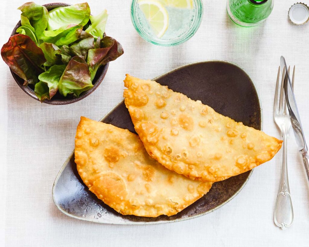 Two chebureki on a dark plate set on a white tablecloth, surrounded by cutlery, glasses and a side salad. Seen from above