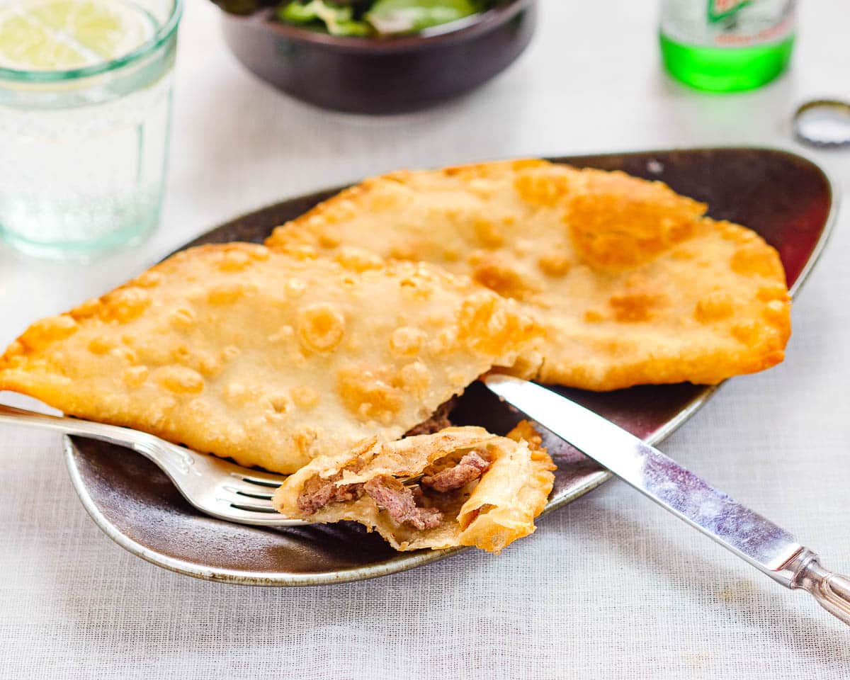 Close-up of a chebureki that has been cut open, with a side salad and glasses in the bakground.