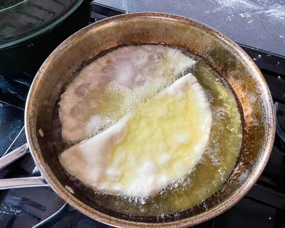 Two cheburekis shallow frying in a pan with high sides.